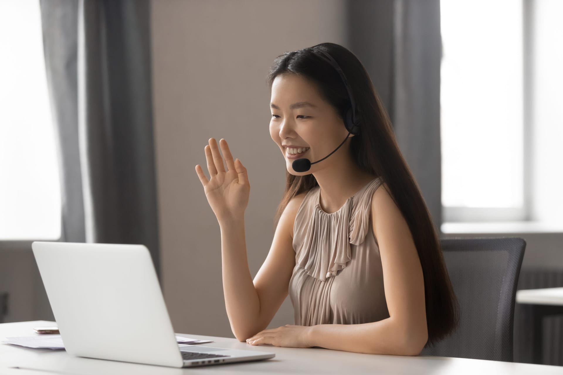 Young Asian woman with headphones and microphone smiling at laptop camera as she interprets languages.